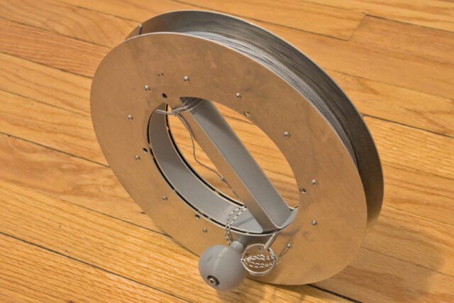 A reel with aluminum sides and a gray plastic handle is wrapped with gray string and propped up on its edge.
