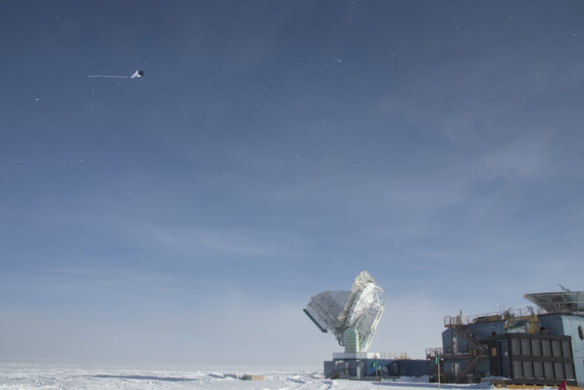 A navy and white kite appears in the top left corner of the image, with its fuzzy tail blown horizontal, to the left. The white 10 meter diameter South Pole Telescope and the blue Dark Sector Laboratory building appear in the bottom right of the photo, with snow along the bottom of the image. Blowing snowflakes are visible against the mostly-clear blue sky.